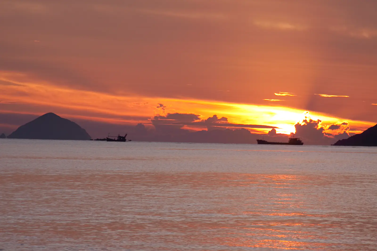 Bình minh trên vịnh Nha Trang: mặt trời ló sau mây, bóng đảo và tàu cá in trên mặt biển cam hồng