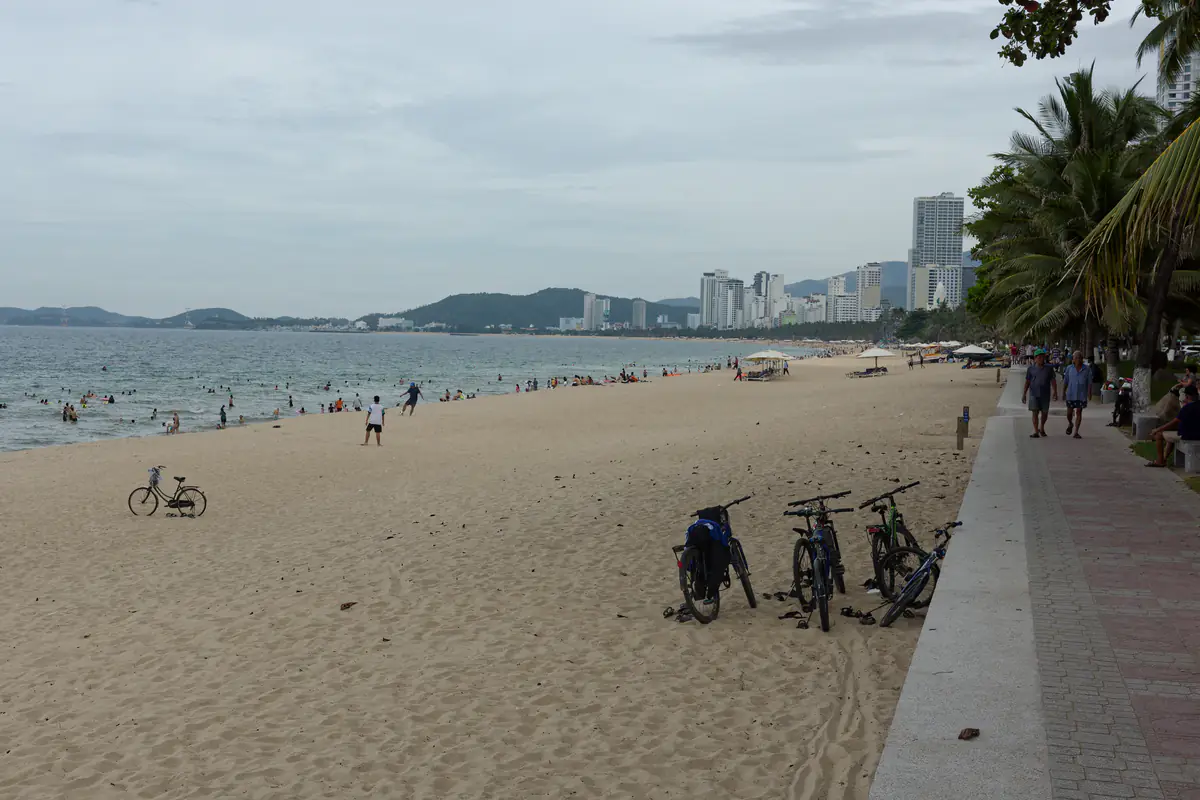Bãi cát đường Trần Phú, Nha Trang buổi chiều nhìn về phía nam, người tắm gần mép nước, xe đạp dựng trên cát, lối đi bộ ven biển và hàng dừa bên phải, tháng 6/2023