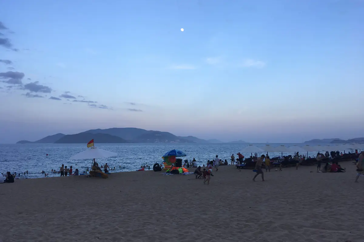 Dù trắng của cứu hộ cắm cờ đỏ vàng, ván cứu hộ màu vàng dưới chân dù trên bãi biển trung tâm Nha Trang lúc chạng vạng, người tắm ở mép nước, phía xa là đảo Hòn Tre, tháng 7/2018