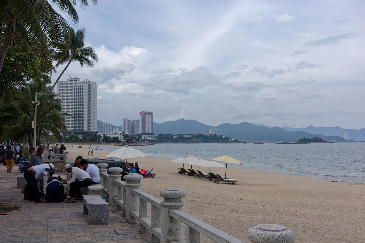 Dãy dù trắng và ghế nằm trên bãi Trần Phú, Nha Trang, bên lan can đá của công viên ven biển, nhìn về phía bắc với núi và nhà cao tầng phía xa, tháng 6/2023