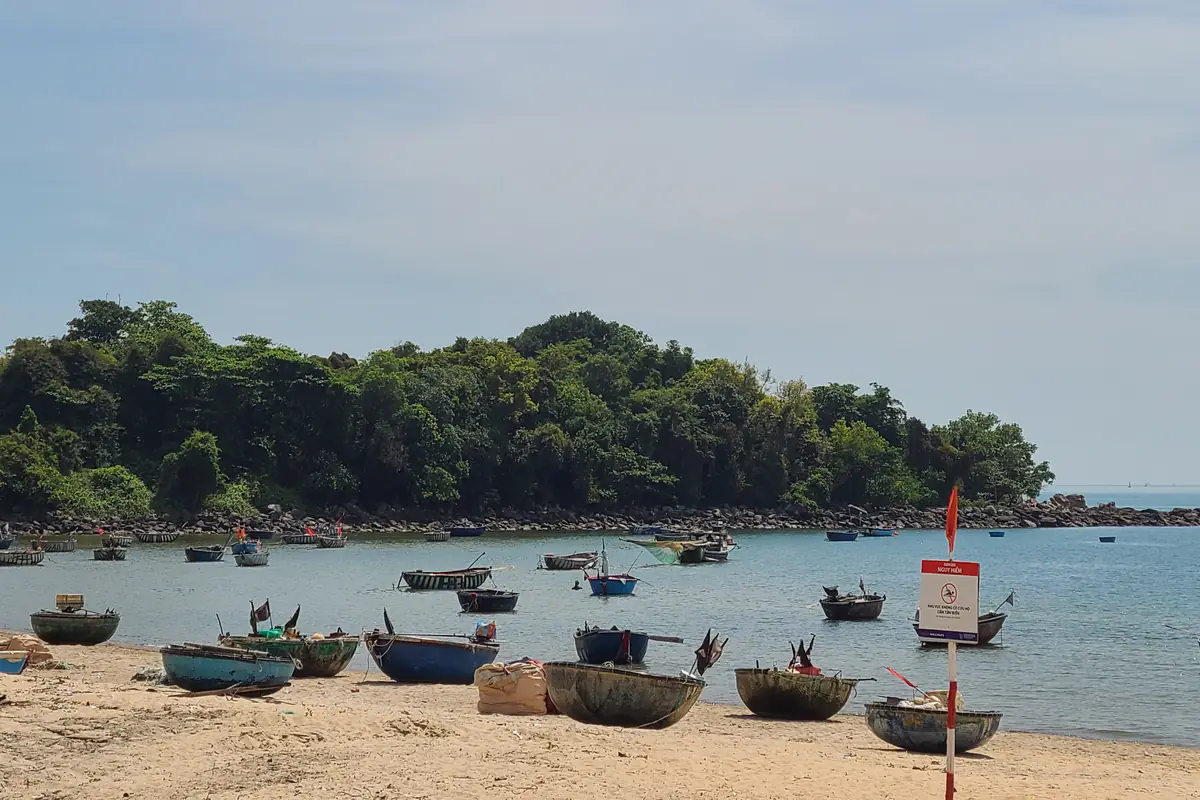 Thuyền thúng neo sát bờ làng chài Nam Ô, Đà Nẵng, mỏm đất phủ cây và rạn đá phía sau, biển cảnh báo cắm trên cát, sáng tháng 5/2025
