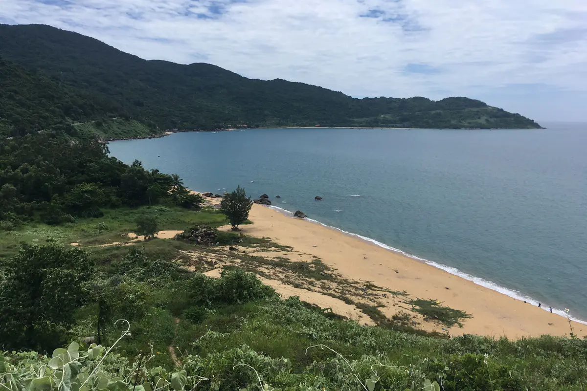 Bờ biển phía nam bán đảo Sơn Trà nhìn từ đường gần chùa Linh Ứng: bãi cát vàng dưới sườn núi phủ rừng, vài tảng đá sát mép nước, tháng 7/2018