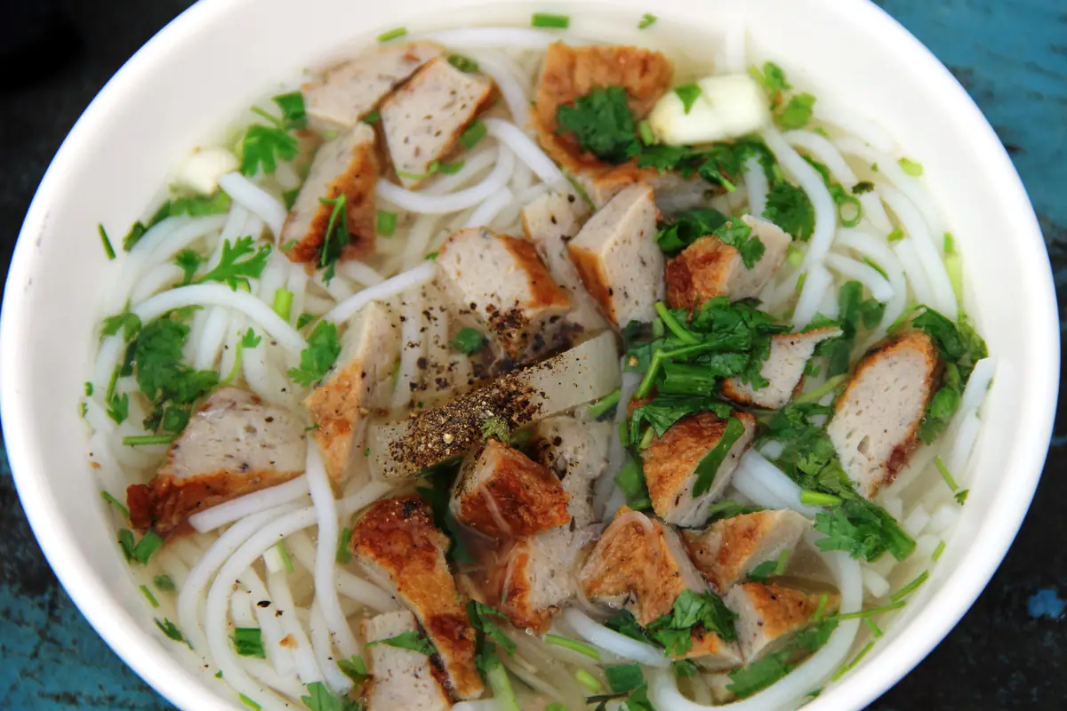 A white bowl of fish cake noodle soup with chunks of golden fried fish cake, rice noodles, cilantro, scallions and cracked black pepper, August 2018