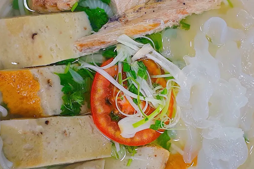 Close-up of a bowl of Nha Trang fish noodle soup: steamed and fried fish cake, fish, white jellyfish and a slice of tomato in broth with herbs and scallions