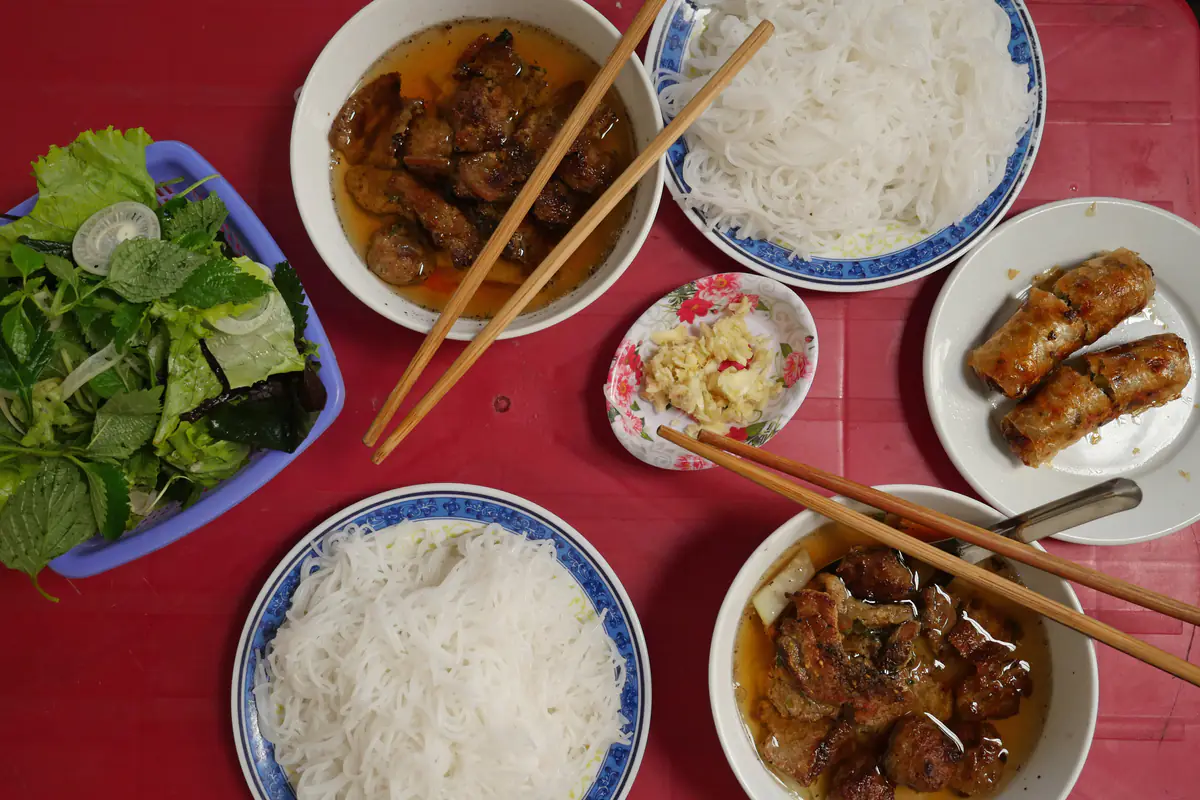 Hai suất bún chả Hà Nội trên bàn nhựa đỏ: hai bát chả nướng ngâm nước chấm màu hổ phách, hai đĩa bún trắng, rổ xà lách và tía tô, đĩa tỏi băm và hai cuốn nem rán, tháng 3/2019