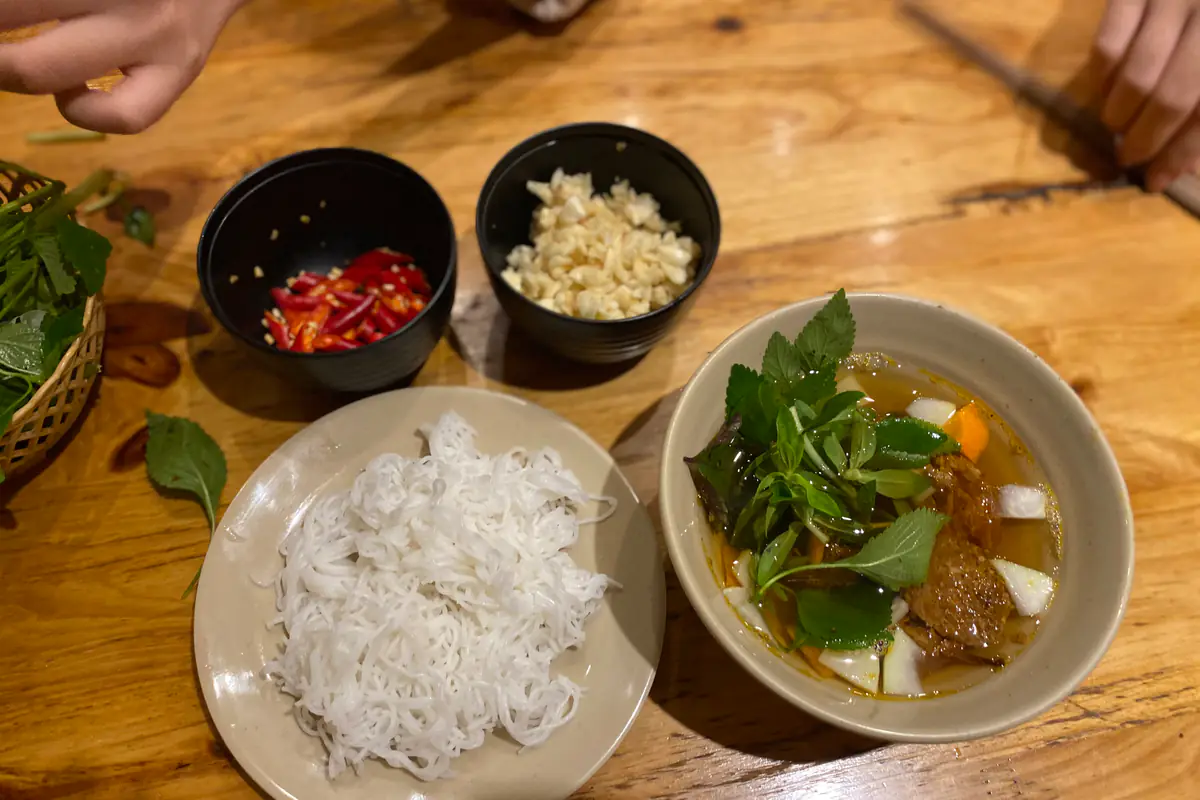 Bát nước chấm bún chả có chả nướng và rau thơm thả thẳng vào bát, bên cạnh đĩa bún, chén ớt tươi thái lát và chén tỏi băm trên bàn gỗ, Hà Nội, tháng 8/2023