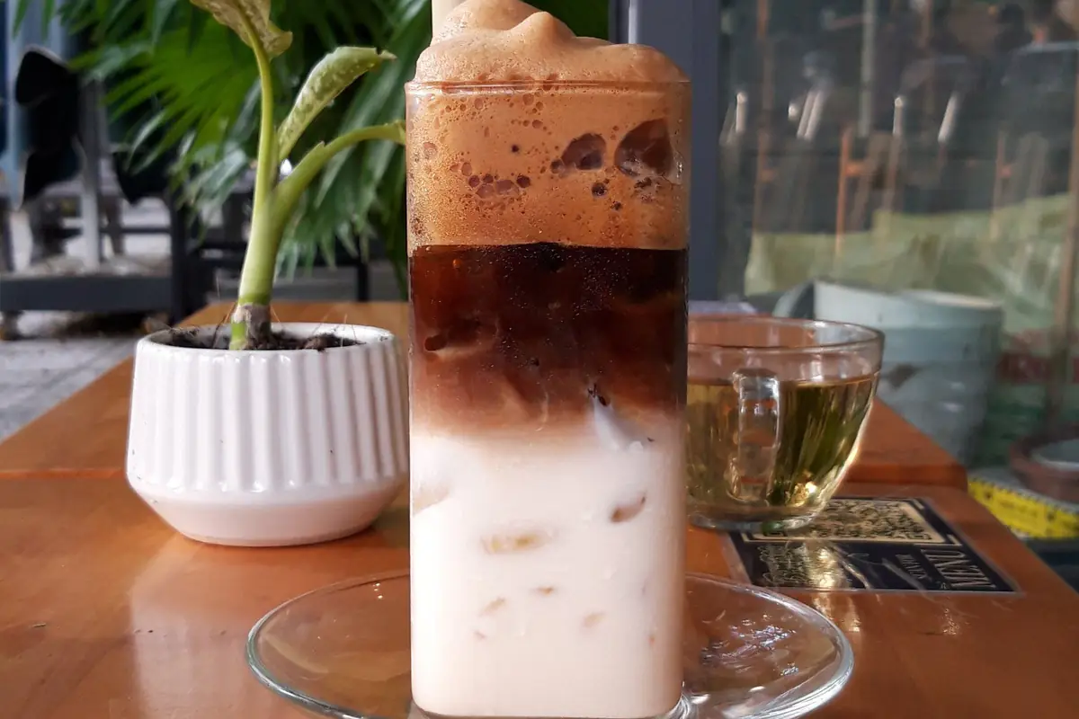Ly bạc xỉu đá cao nhiều lớp: sữa trắng có đá ở dưới, lớp cà phê nâu sẫm ở giữa, lớp bọt cà phê trên cùng, ống hút giấy, đặt trên đĩa thủy tinh cạnh chậu cây nhỏ và tách trà trong một quán ở Đà Nẵng, tháng 11/2023