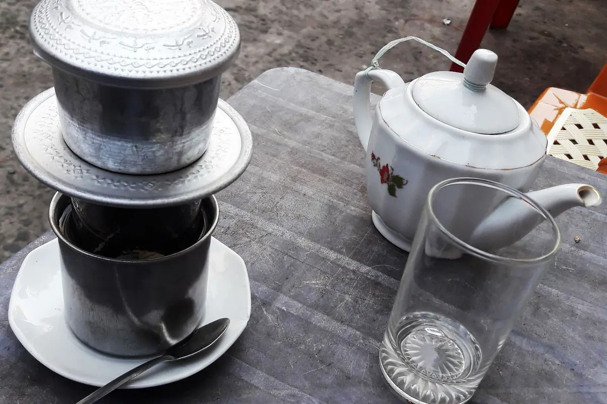 Phin cà phê nhôm đặt trên cốc inox và đĩa sứ, bên cạnh ấm trà sứ hoa hồng và ly thủy tinh rỗng trên bàn nhựa xám của một quán vỉa hè, ghế nhựa cam phía sau, quận 3, TP.HCM, tháng 3/2020