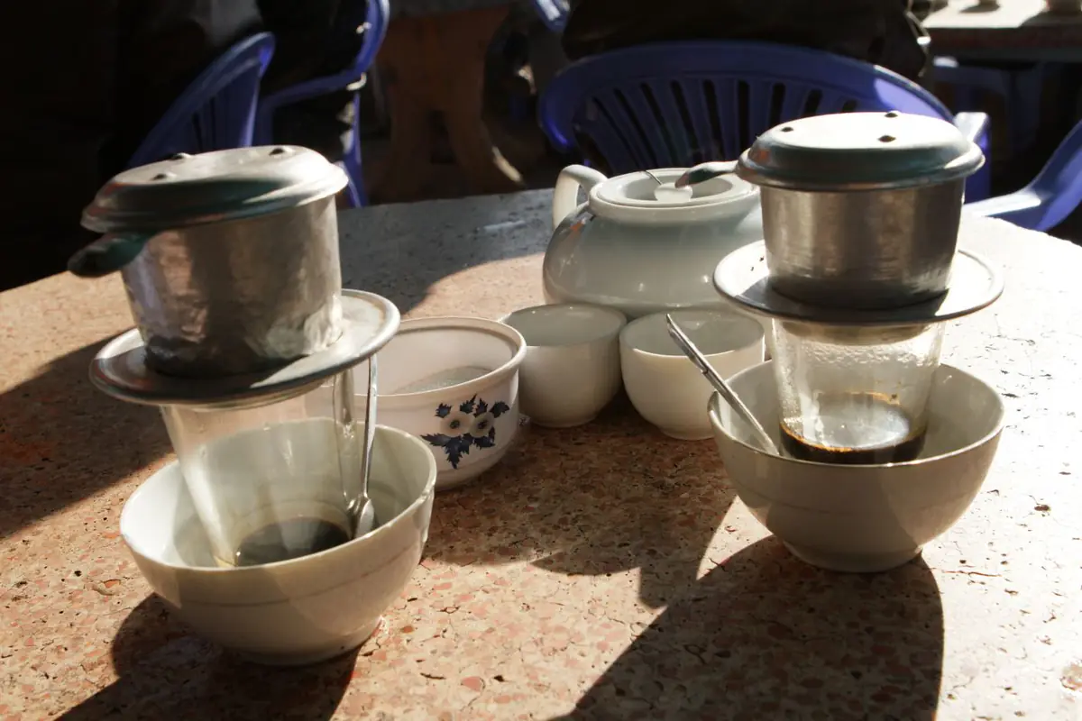 Hai phin cà phê nhôm đặt trên ly thủy tinh, mỗi ly ngồi trong một bát nước nóng, bên cạnh ấm trà, chén trà và hũ đường trên bàn đá mài của một quán cà phê ở Đà Lạt, ghế nhựa xanh phía sau, tháng 2/2015