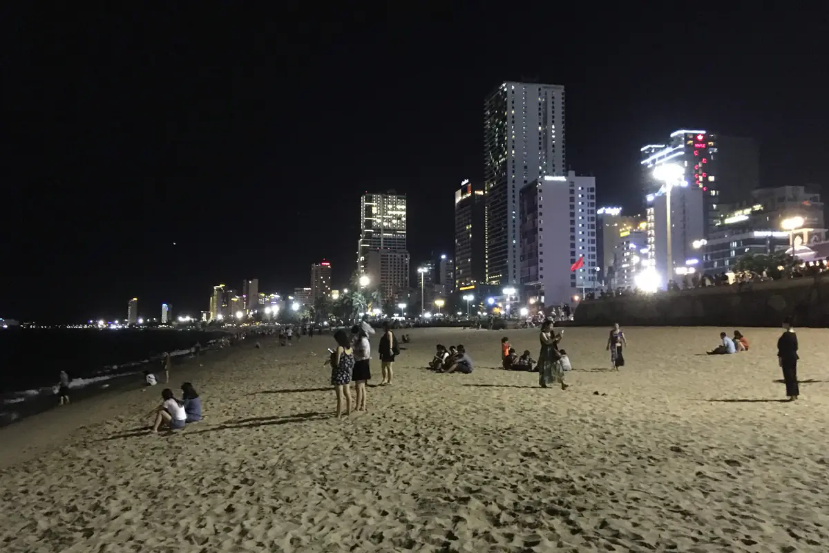 Người dân và du khách ngồi hóng gió trên bãi cát Trần Phú buổi tối, phía sau là dãy khách sạn cao tầng lên đèn