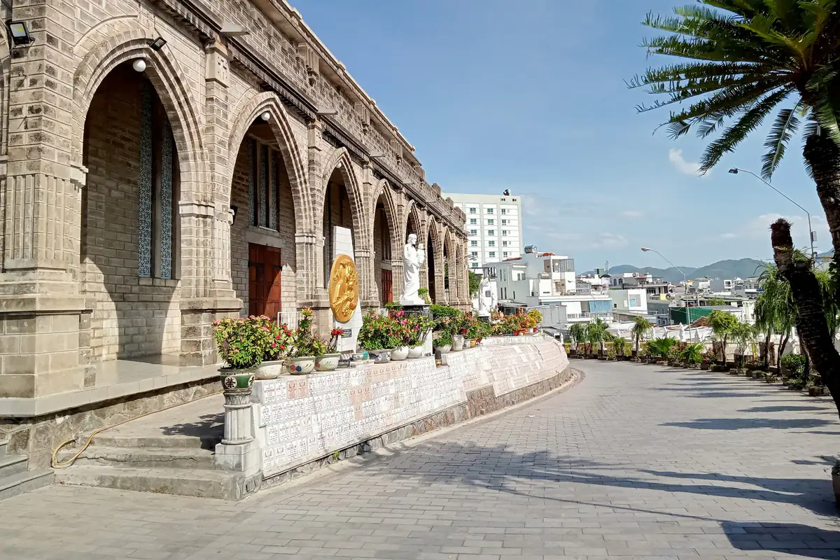 Dãy vòm đá kiểu Gothic dọc lối vào lát gạch của Nhà thờ Núi Nha Trang, chậu hoa và tượng trắng bên tường, phía xa là phố và núi, nắng chiều tháng 7/2022