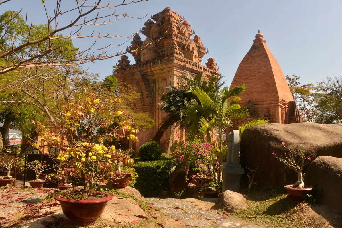 Tháp chính và một ngọn tháp mái nhọn bằng gạch ở Tháp Bà Ponagar trong nắng sáng, phía trước là chậu mai vàng, cây cau và tảng đá lớn, tháng 2/2014