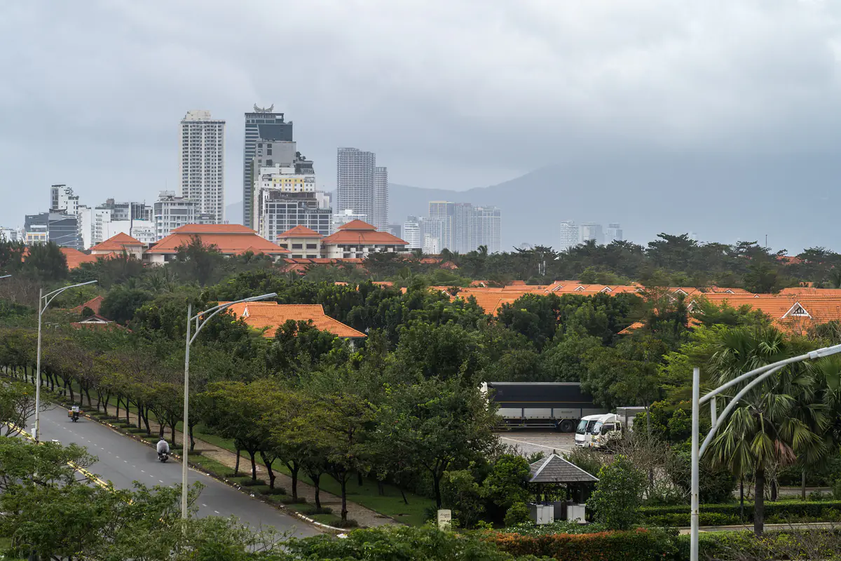 Nhà cao tầng Đà Nẵng trưa 26/11/2024 dưới mây xám thấp, núi phía xa chìm trong mây, phía trước là hàng cây xanh và mái ngói đỏ