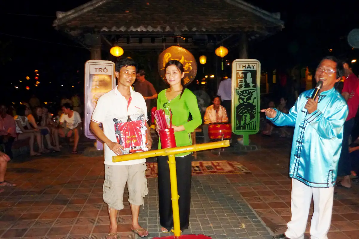 Anh hiệu bài chòi mặc áo xanh cầm micro giới thiệu hai người chơi trúng thưởng ở sân bài chòi phố cổ Hội An, chòi mái ngói và đèn lồng phía sau, khán giả ngồi quanh, tháng 5/2012