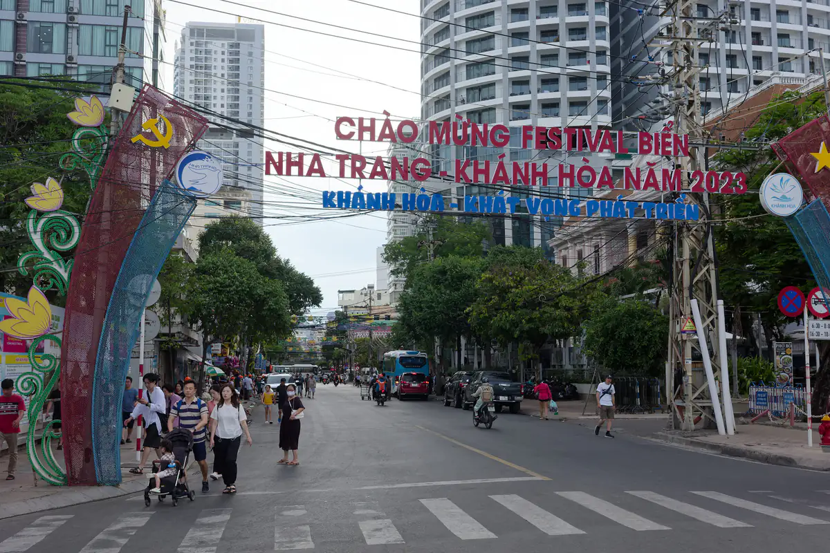 Người đi bộ qua vạch kẻ đường trên phố Nguyễn Thị Minh Khai, Nha Trang, cổng chào Festival Biển 2023, xe buýt và xe máy phía sau, tháng 6/2023