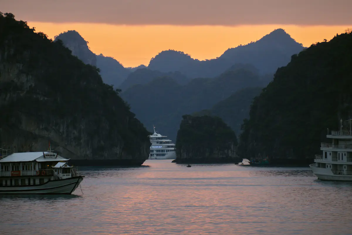 Tàu du lịch và du thuyền neo giữa các đảo đá vôi trên vịnh Hạ Long lúc chạng vạng, trời ngả màu cam phía sau dãy núi
