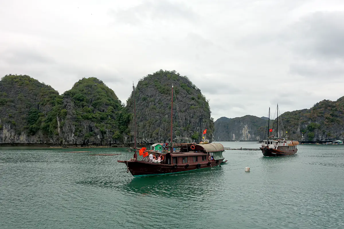 Hai thuyền gỗ chở khách chạy trên vịnh Lan Hạ giữa các đảo đá vôi phủ cây, trời nhiều mây, tháng 4/2019