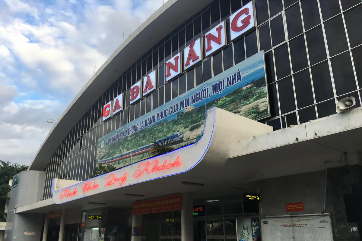 Mặt tiền ga Đà Nẵng với dòng chữ Ga Đà Nẵng trên mái cong, biển Kính chào quý khách và bảng giờ tàu ở lối vào, buổi sáng tháng 7/2018
