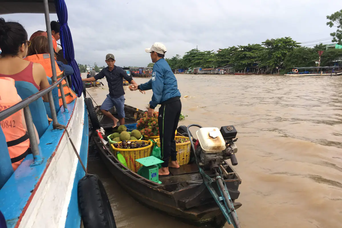Du khách mặc áo phao cam ngồi trên tàu, người bán trên ghe nhỏ chở mít và dừa chuyền trái cây sang ở chợ nổi Cái Răng, tháng 7/2018