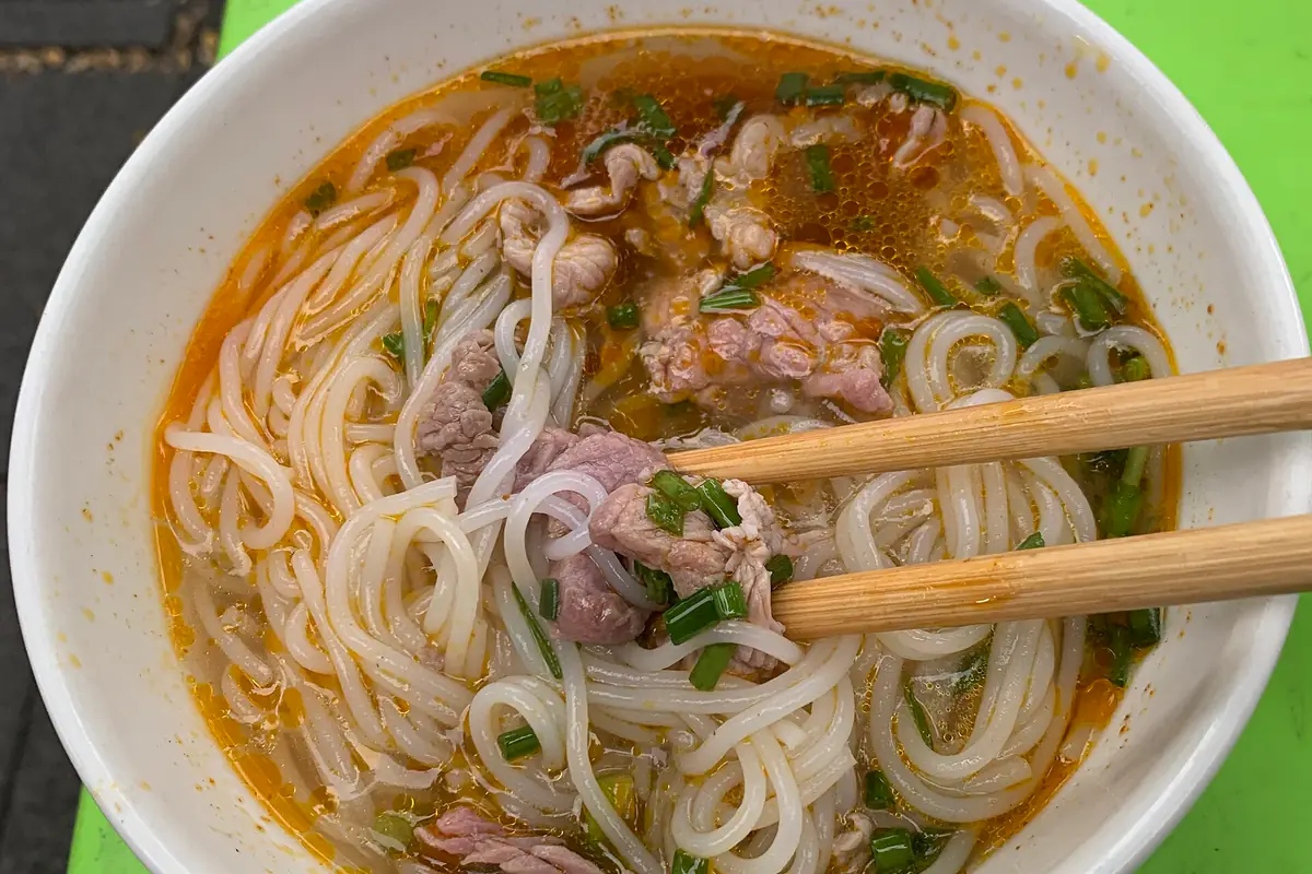 Tô bún bò Huế ở chợ Đông Ba, Huế: sợi bún to, thịt bò, hành lá trong nước dùng màu đỏ cam có dầu ớt, đôi đũa gỗ gắp sợi bún, tháng 7/2024