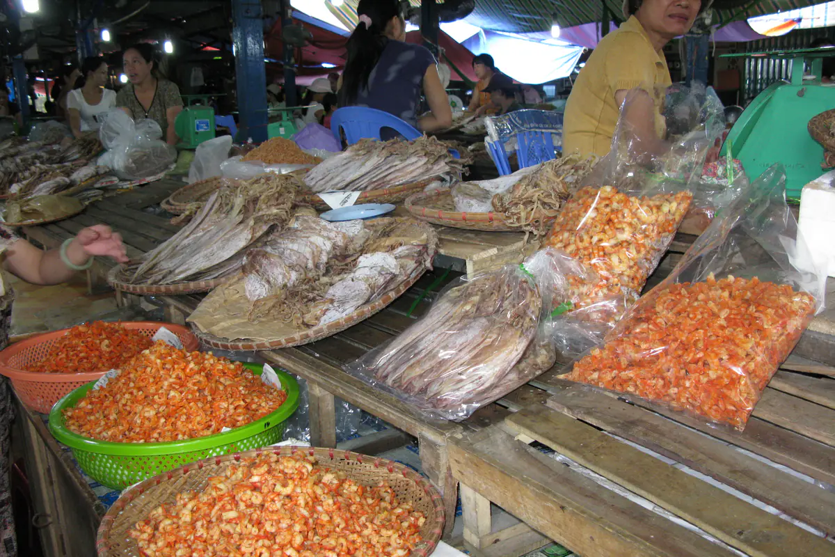 Sạp đồ khô trong chợ Cát Bà bày mực khô trên nia và tôm khô trong rổ, túi nilon, người bán ngồi phía sau, tháng 8/2008