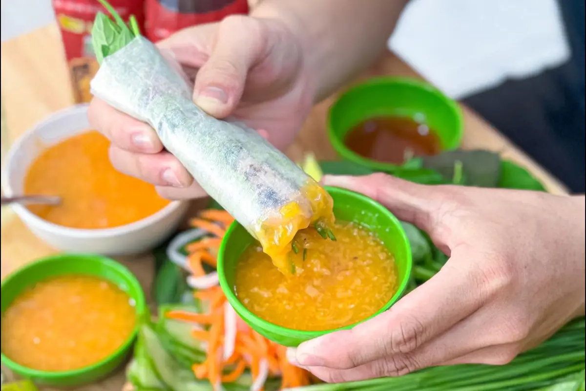 Bàn tay chấm một cuốn nem nướng vào chén nước chấm sệt màu cam, xung quanh là thêm vài chén nước chấm, chén nước mắm, rau sống và đồ chua
