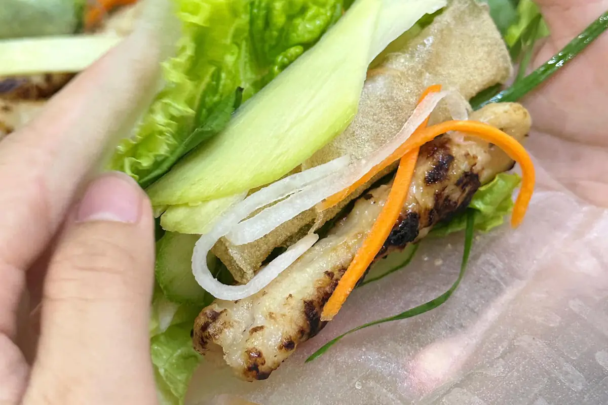 Hai bàn tay đang cuốn nem nướng trên lá bánh tráng mềm, bên trong có xà lách, lát xoài xanh, ram giòn, đồ chua và một cây nem nướng đã rút que
