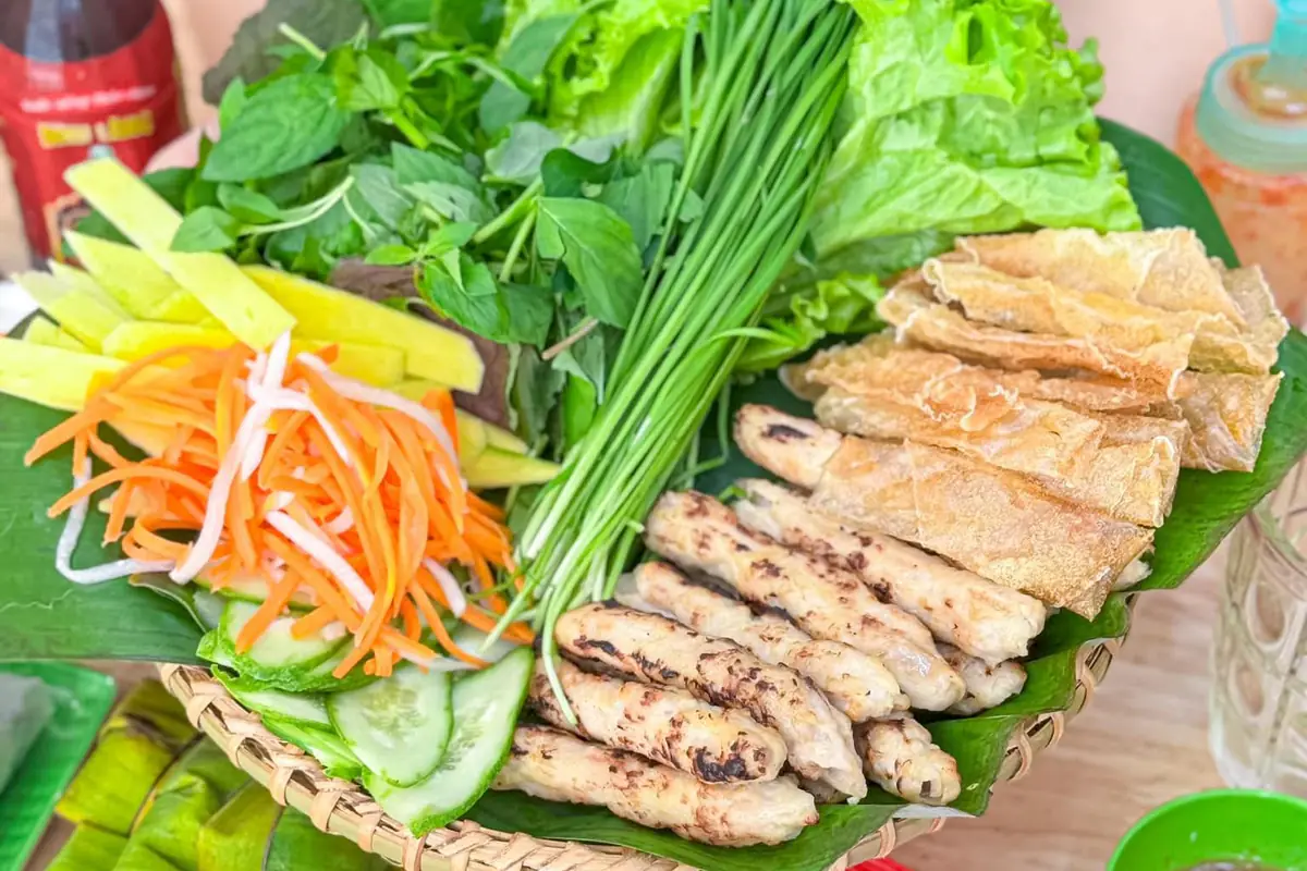 Cận cảnh mẹt nem nướng: xiên nem nướng xém cạnh, ram giòn, xà lách, rau thơm, bó hẹ, dưa leo, đồ chua cà rốt củ cải và xoài xanh xếp trên lá chuối