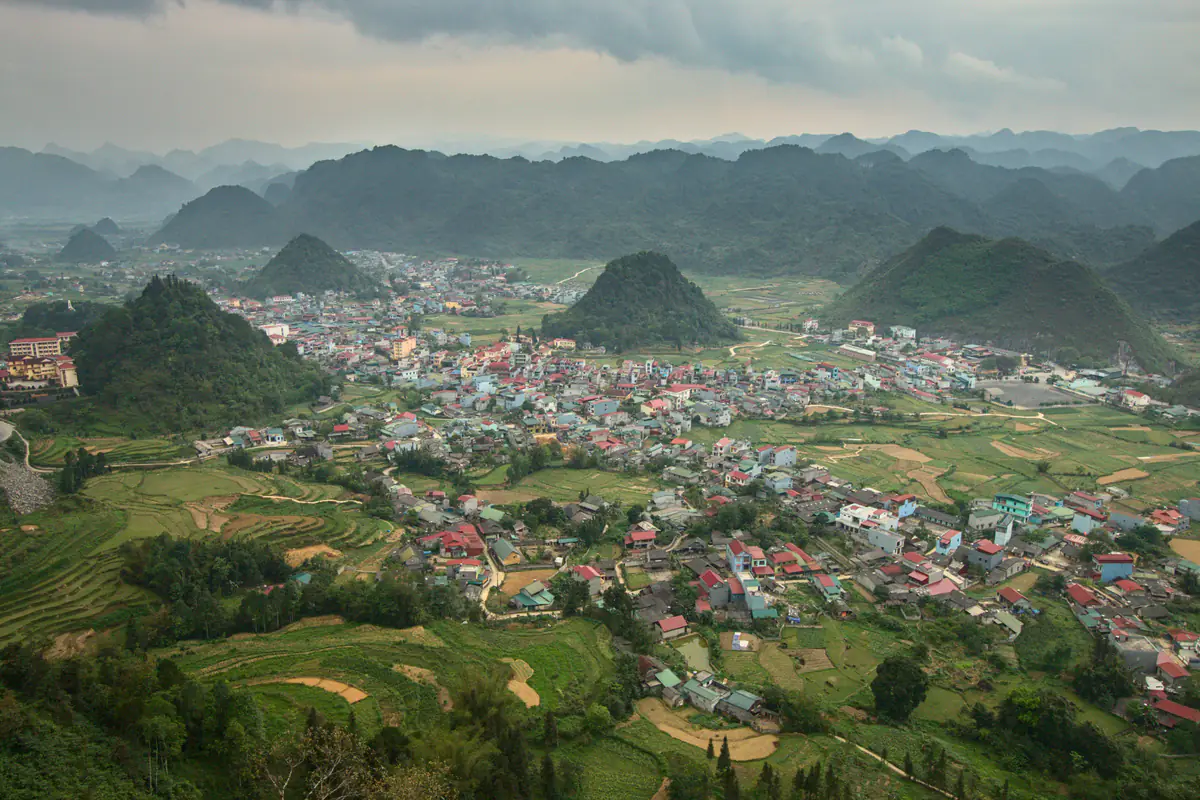 Thung lũng Quản Bạ nhìn từ trên cao: khu nhà mái đỏ, xanh giữa ruộng và những quả đồi tròn, dãy núi đá vôi phía sau dưới trời nhiều mây, tháng 4/2023