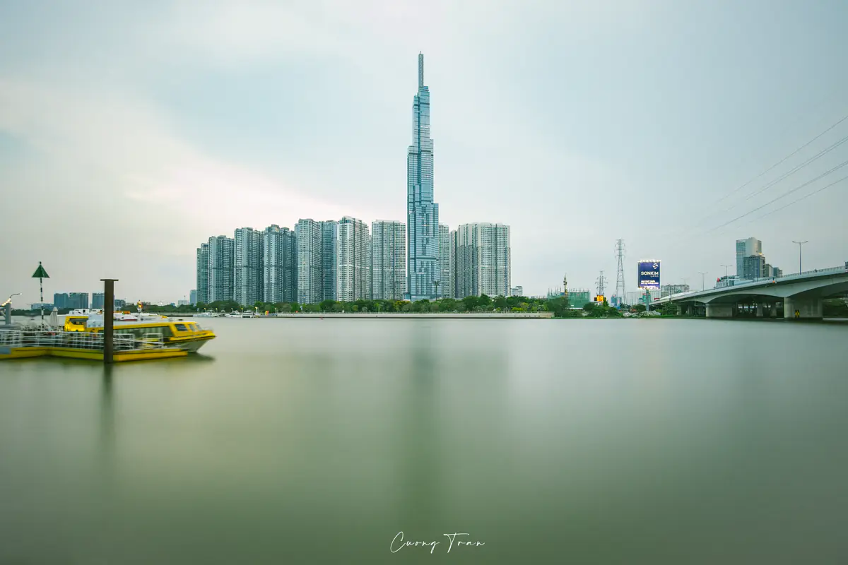 Tàu buýt sông màu vàng xanh trên sông Sài Gòn, phía sau là tòa Landmark 81 và dãy chung cư ven sông, trời nhiều mây, tháng 4/2022