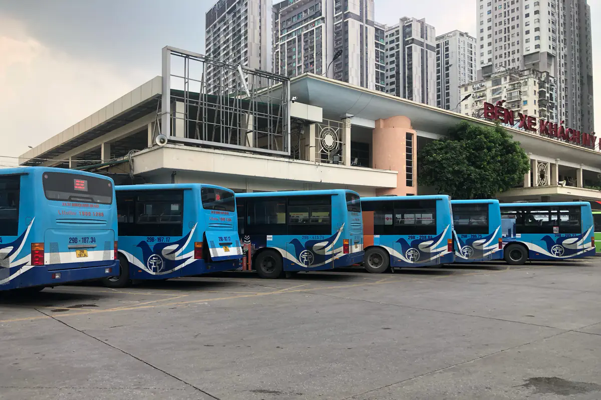 Một hàng xe buýt xanh trắng của Hà Nội đỗ ở bến xe Mỹ Đình, phía sau là nhà ga bến xe và các tòa chung cư cao tầng, tháng 7/2025