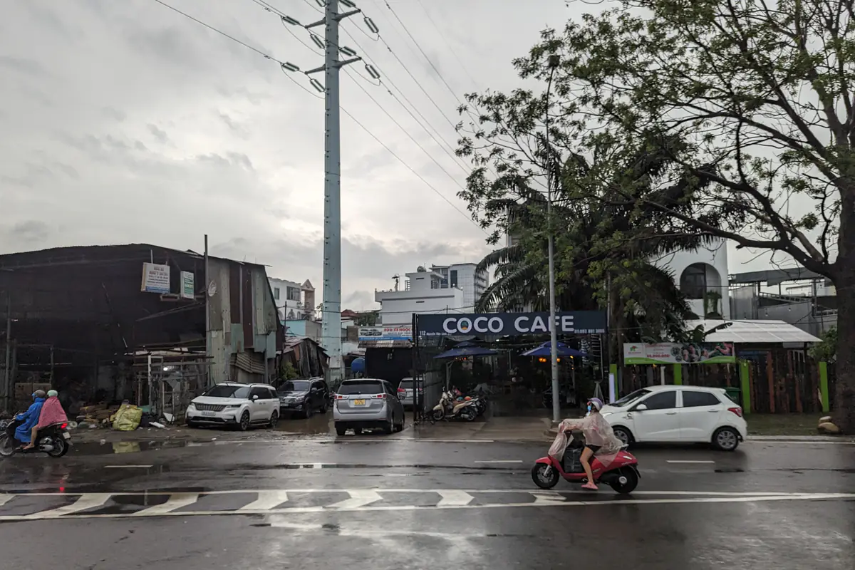 Mặt đường ướt loáng sau mưa, người đi xe máy mặc áo mưa trong suốt, xe ô tô đỗ trước dãy quán, trời nhiều mây xám, khu quận 7 cũ TP.HCM, chiều 1/7/2023