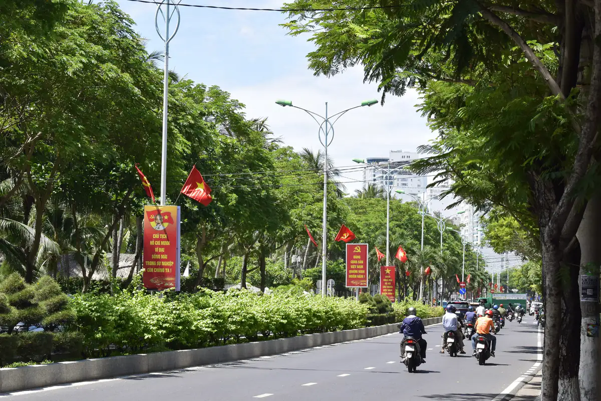 Người đi xe máy đội mũ bảo hiểm chạy trên một đại lộ rợp cây ở Nha Trang, dải phân cách trồng cây bụi và cắm cờ, buổi sáng tháng 6/2025