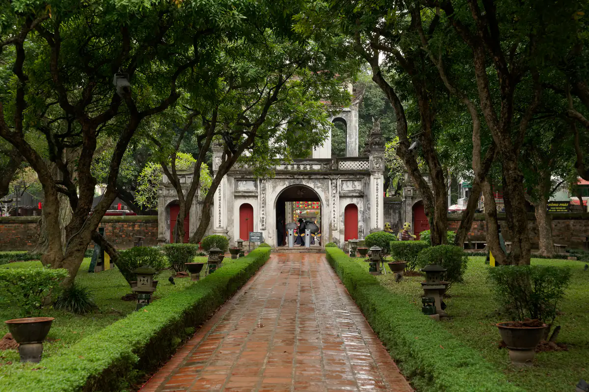 Lối gạch đỏ ướt mưa giữa hai hàng cây cổ thụ và thảm cỏ trong khu Nhập Đạo, cuối lối là cổng Văn Miếu Môn ba cửa, Văn Miếu - Quốc Tử Giám, tháng 1/2024